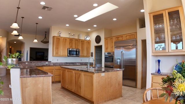 kitchen featuring hanging light fixtures, stainless steel appliances, an island with sink, kitchen peninsula, and dark stone counters