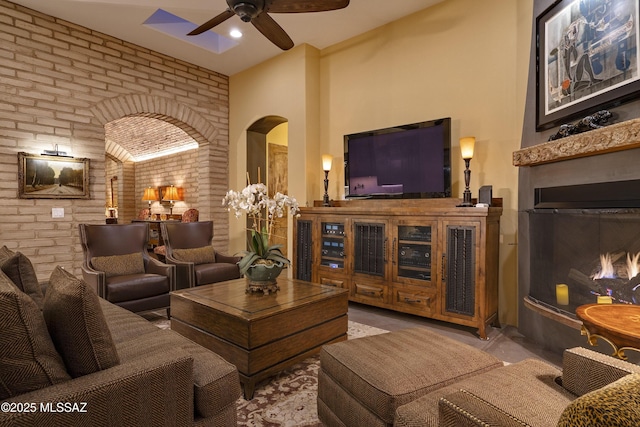 living area with arched walkways, a warm lit fireplace, ceiling fan, and brick wall