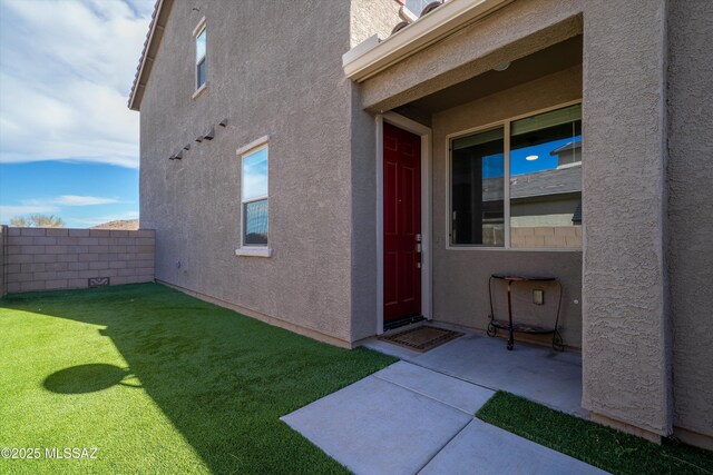 view of exterior entry with a patio, fence, a lawn, and stucco siding