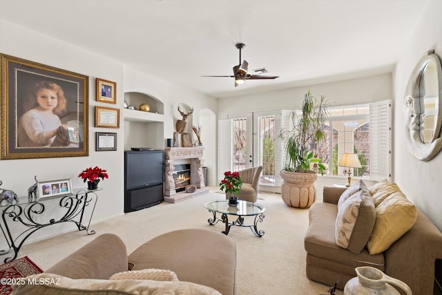 living room featuring built in features, a ceiling fan, a glass covered fireplace, light colored carpet, and french doors
