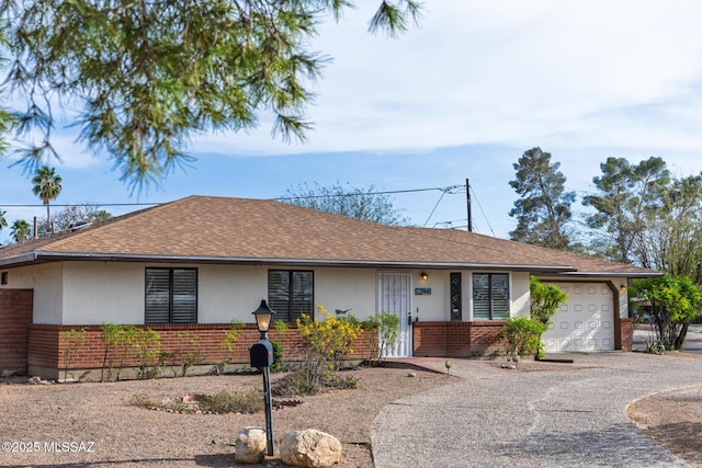 single story home with driveway, a garage, brick siding, roof with shingles, and stucco siding