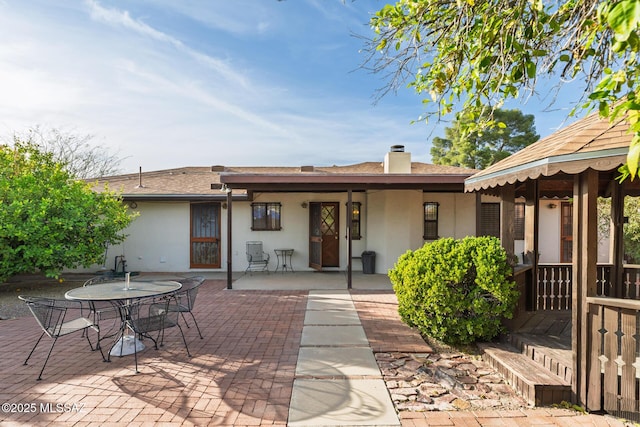 rear view of property featuring stucco siding, roof with shingles, outdoor dining space, a patio area, and a chimney