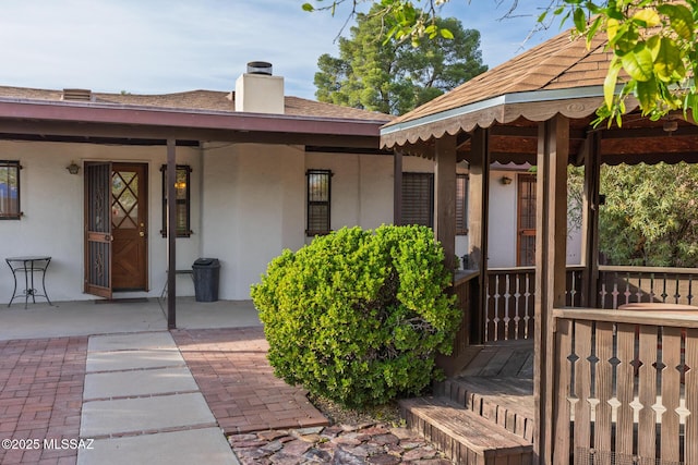 doorway to property with covered porch, stucco siding, roof with shingles, and a chimney