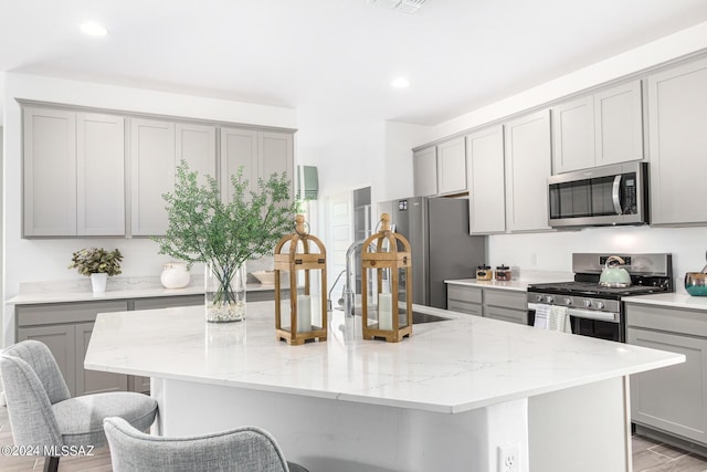 kitchen with sink, gray cabinets, stainless steel appliances, light stone counters, and an island with sink