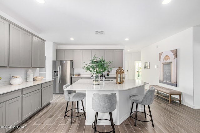 kitchen featuring gray cabinetry, stainless steel fridge with ice dispenser, an island with sink, and a kitchen bar