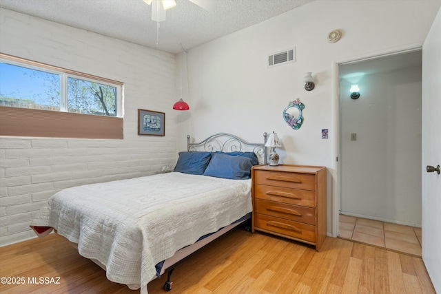 bedroom with visible vents, brick wall, light wood-style flooring, a textured ceiling, and a ceiling fan