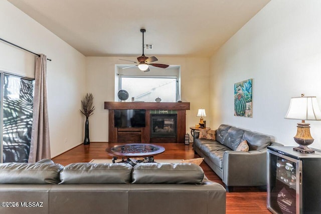 living area with beverage cooler, visible vents, a ceiling fan, a glass covered fireplace, and wood finished floors