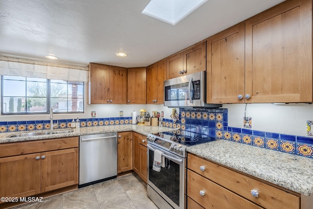 kitchen with light stone counters, a skylight, brown cabinets, appliances with stainless steel finishes, and a sink