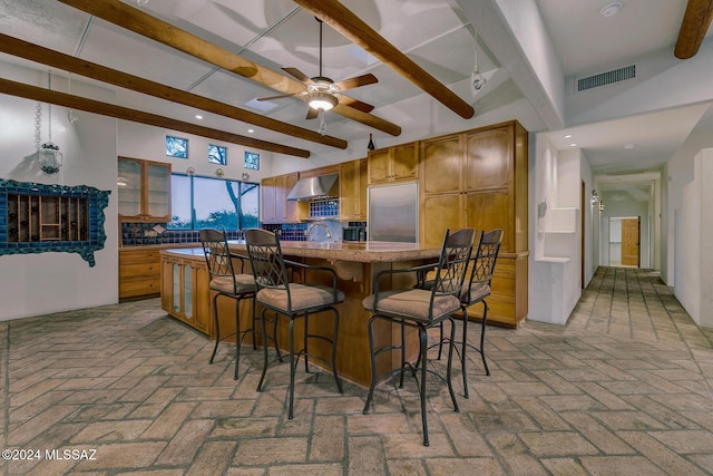 kitchen featuring wall chimney range hood, backsplash, a kitchen bar, stainless steel built in fridge, and beamed ceiling