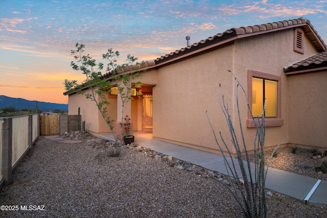 property exterior at dusk with a mountain view, a tile roof, fence, stucco siding, and a gate
