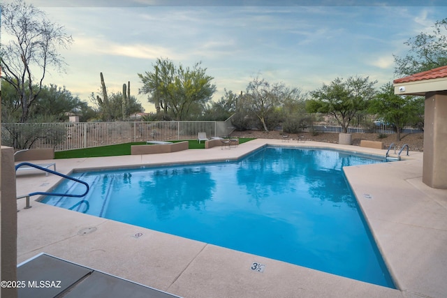 pool at dusk with a patio area, fence, and a community pool