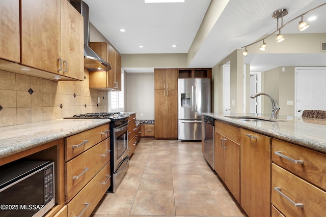 kitchen featuring light stone countertops, stainless steel appliances, a sink, brown cabinets, and wall chimney exhaust hood