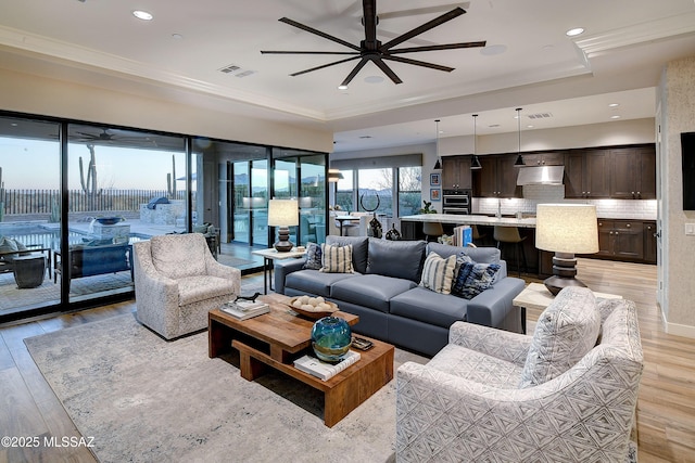 living room with visible vents, a ceiling fan, light wood-type flooring, a raised ceiling, and crown molding