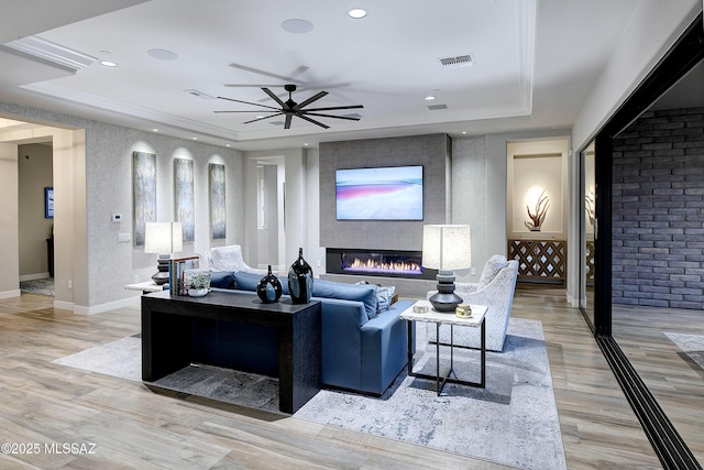 living room with a large fireplace, light wood finished floors, visible vents, a raised ceiling, and ceiling fan