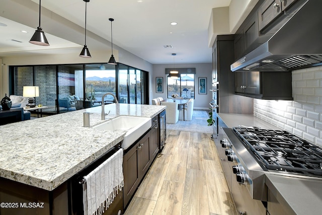 kitchen with a center island with sink, wall chimney exhaust hood, hanging light fixtures, stainless steel gas cooktop, and a sink