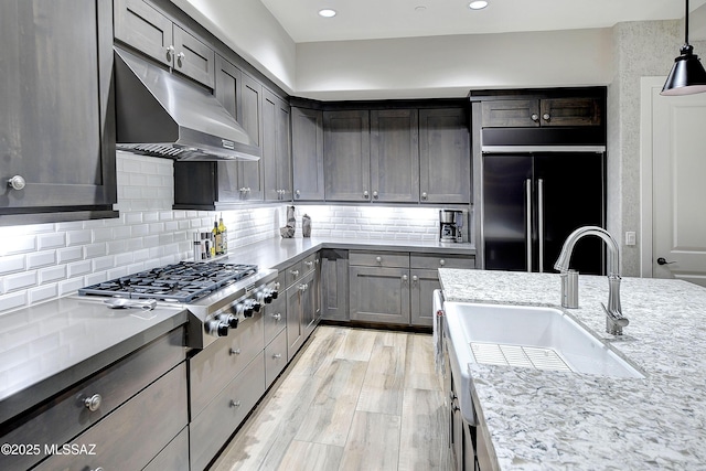 kitchen with light stone counters, high end black fridge, under cabinet range hood, stainless steel gas cooktop, and a sink