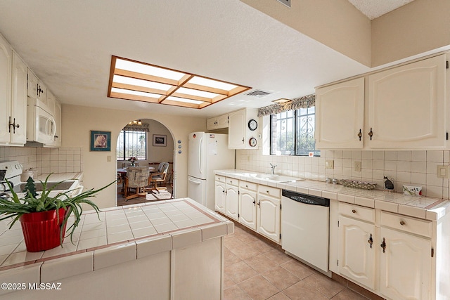 kitchen with arched walkways, tile counters, a sink, white appliances, and a peninsula