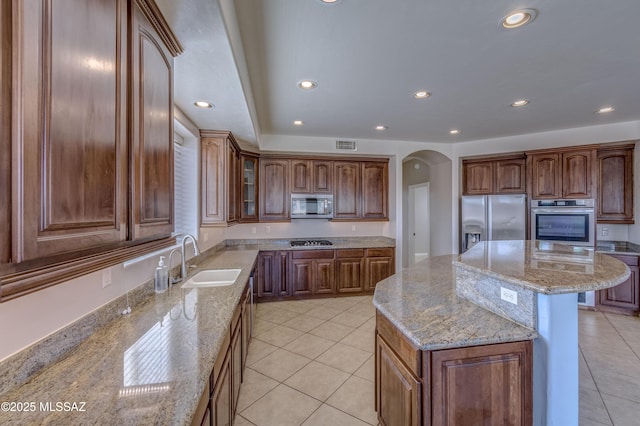 kitchen featuring arched walkways, stainless steel appliances, a kitchen island, light stone countertops, and brown cabinetry