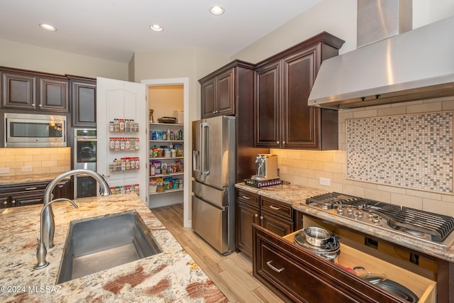 kitchen with a sink, wall chimney range hood, light stone counters, appliances with stainless steel finishes, and light wood-style floors