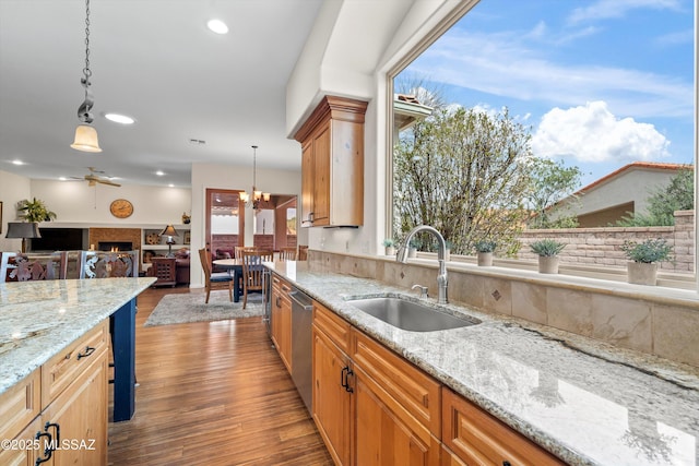 kitchen with wood finished floors, stainless steel dishwasher, a sink, light stone countertops, and a warm lit fireplace