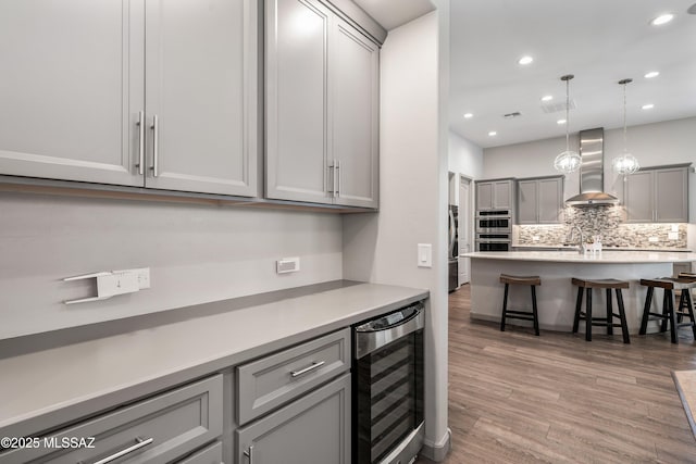 kitchen with light wood-style flooring, gray cabinets, decorative backsplash, wine cooler, and wall chimney range hood