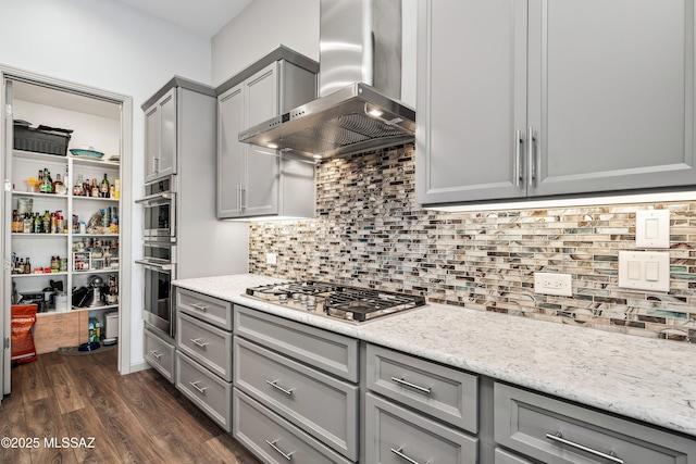 kitchen featuring gray cabinetry, dark wood-type flooring, wall chimney range hood, decorative backsplash, and stainless steel gas stovetop