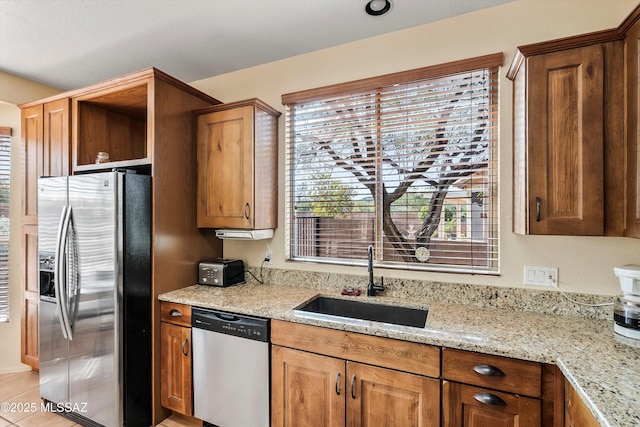 kitchen with brown cabinetry, light stone counters, stainless steel appliances, and a sink