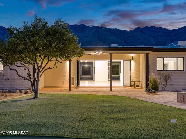 back of house at dusk featuring a mountain view, a lawn, stucco siding, and a patio area