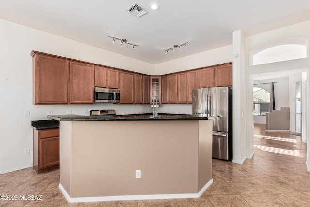 kitchen with stainless steel appliances, brown cabinetry, a center island with sink, and glass insert cabinets