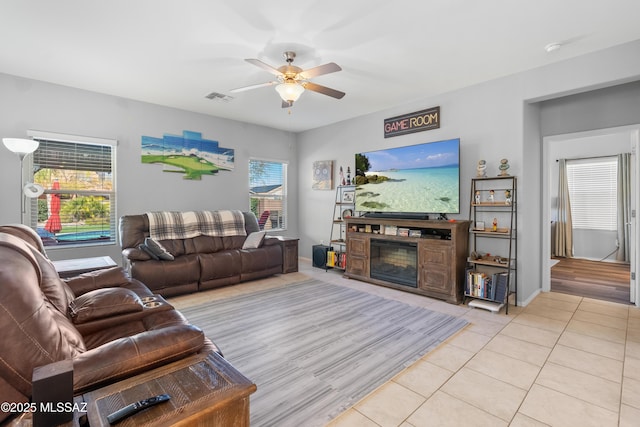 living room featuring light tile patterned floors, ceiling fan, visible vents, baseboards, and a glass covered fireplace