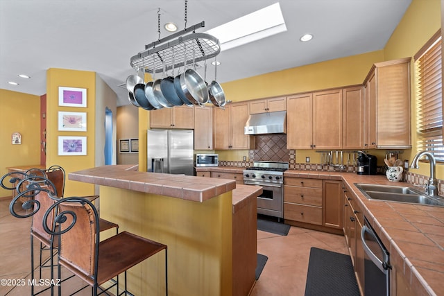 kitchen featuring tile counters, appliances with stainless steel finishes, a sink, a kitchen island, and under cabinet range hood