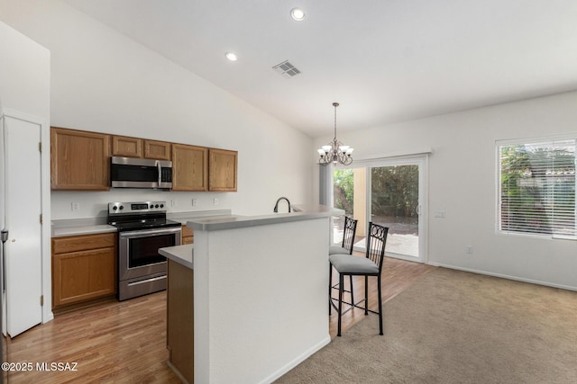 kitchen featuring pendant lighting, light countertops, appliances with stainless steel finishes, brown cabinetry, and a kitchen island with sink