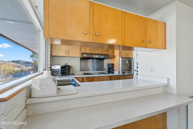 kitchen with wall oven, brown cabinetry, light countertops, under cabinet range hood, and gas cooktop