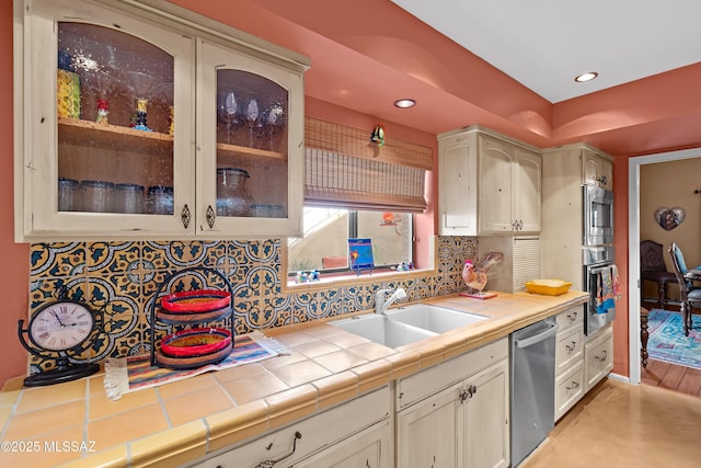 kitchen with tile countertops, stainless steel appliances, backsplash, glass insert cabinets, and a sink