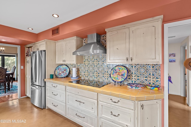 kitchen with tile counters, freestanding refrigerator, black electric stovetop, wall chimney range hood, and backsplash