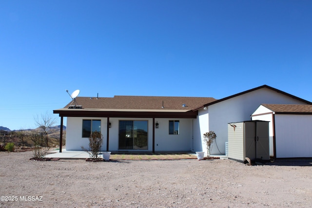 back of house featuring an outbuilding, a shingled roof, stucco siding, a shed, and a patio area