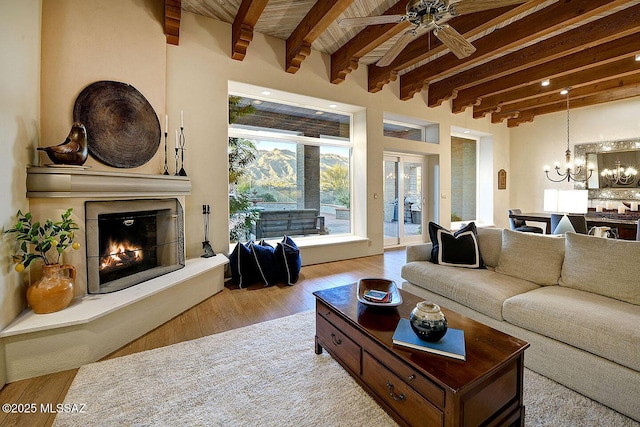 living room with a lit fireplace, ceiling fan with notable chandelier, beam ceiling, and wood finished floors