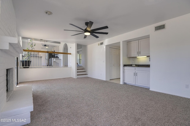 unfurnished living room featuring a fireplace, visible vents, light colored carpet, stairway, and a ceiling fan