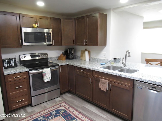 kitchen with light wood finished floors, light stone counters, stainless steel appliances, dark brown cabinets, and a sink