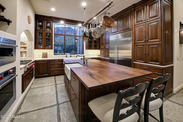 kitchen featuring a kitchen island with sink, decorative backsplash, stainless steel appliances, and wooden counters