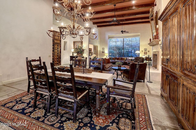 dining area featuring a fireplace, a towering ceiling, beamed ceiling, baseboards, and ceiling fan with notable chandelier