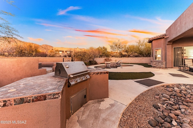 patio terrace at dusk featuring a fire pit, an outdoor kitchen, a grill, and fence