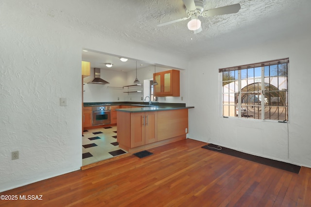 kitchen featuring pendant lighting, brown cabinets, dark countertops, wall chimney range hood, and oven