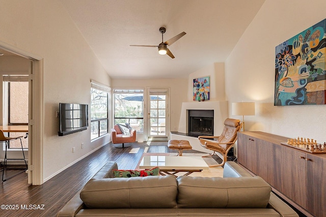 living room featuring dark wood-style flooring, a fireplace with raised hearth, vaulted ceiling, ceiling fan, and baseboards