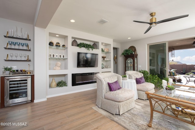 living area with built in shelves, wine cooler, light wood finished floors, a dry bar, and a glass covered fireplace