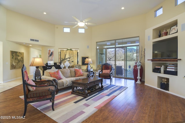 living area with visible vents, a towering ceiling, hardwood / wood-style floors, a ceiling fan, and baseboards