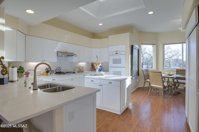 kitchen with double oven, under cabinet range hood, stainless steel gas cooktop, a sink, and light wood finished floors