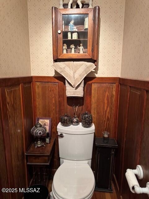 half bathroom with wainscoting, toilet, and wallpapered walls