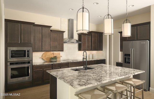 kitchen with dark brown cabinetry, light stone countertops, a sink, wall chimney exhaust hood, and stainless steel appliances