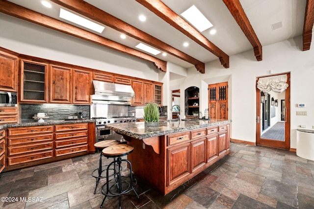 kitchen featuring glass insert cabinets, range hood, a kitchen island with sink, and stone tile floors